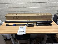 Top view of the Vivo projector screen components laid out on a wooden desk including tripod, screen frame, support rod, instruction manual, and original box.