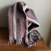 Grey wool knit scarf with horizontal stripes in bright blue and crimson displayed draped over a brown surface, showing texture and fringe ends.