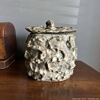 Front view of Brutalist volcanic glazed pottery lidded vessel showing thick textured lava glaze and loop handle on lid.