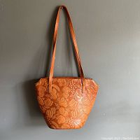 Full front view of the medium brown hand tooled leather handbag with long shoulder straps against a gray background.
