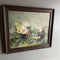 Framed oil painting showing garden pansies in a cluster with greenery, hung on a wall.