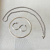 Sterling silver twisted rope chain necklace, etched bangle bracelet, and half hoop earrings laid out on fabric background.