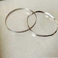 Two sterling silver bangles shown together on a light fabric background, one with wave shape and one with Greek key cutout