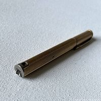 Full side view of the vintage Colibri pen lighter lying flat on a fabric surface, showing its gold-tone finish and clip.