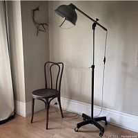 Full view of the surgical floor lamp standing beside a wooden chair, showing the black base on casters, tall adjustable arm, and tilted lamp head.