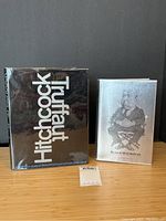 Two books side by side on wooden surface against black background: black dust jacket and silver cloth boards