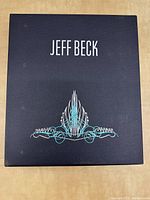 Black linen slipcase with 'Jeff Beck' title in white above turquoise stylized emblem.