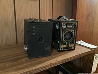Two vintage Brownie box cameras side by side on a wooden shelf, showing front and side profiles.