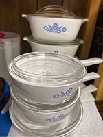 Stacked white Corning Ware Blue Cornflower casseroles with glass lids on top, showing blue floral pattern and clear lids.
