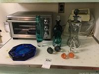 Nine collectible glass pieces on a kitchen counter: two decanters with stoppers, one teal and one clear; a dark green glass cat figurine; blue octagonal ashtray; two orange and two gray bird glass figurines.