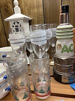 Photo showing multiple glasses with white label text and Christmas tree design, alongside white wooden birdhouse and Jim Beam spark plug decanter.