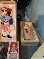 Faith Wick Clown doll lying in an open box with white tissue paper, dressed in colorful clown attire with red hat, blue and white striped shirt and gloves; Pierrot doll in box with clear visor to right; Ideal Baby Baby doll in original packaging below.