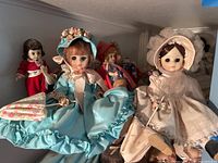 Photo showing three seated Madame Alexander dolls in elaborate fabric costumes including a blue dress with floral bonnet, white lace bonnet and dress, and red costume with white sash, displayed on a shelf.
