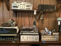 Wide view of shelf and table displaying all components: Fisher unit on top left, Technics tuner on top right, Magnavox and Technics turntables and receivers below, carton of cords on right