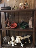 Three shelves showing multiple figurines including ceramic cats, piggy bank, glass bowl and other decorative items.