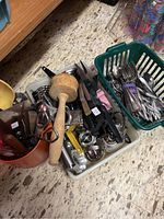 Overview photo showing assorted kitchen utensils and flatware including wooden mallet, graters, can opener, and scoops gathered in containers on floor.