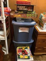 Photo of stacked board games including Super Simon, Othello, Po-Ke-No card decks, and two blue totes labeled for jigsaw puzzles, with toys and crayons on top.