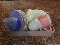 Set of pastel-colored cups, saucers, and plates stacked in a box on carpet.