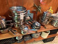 Wide view of multiple stainless steel pots, pans, muffin tins, cutting boards and ceramic crock on wooden shelving.
