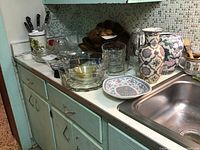 Kitchen counter showing multiple trinket boxes, ceramic vases, and Pyrex glass containers arranged near the sink beneath mosaic tile backsplash.