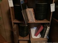 Two shelves with stacks of approximately 45 RPM records and empty jackets, wooden shelving unit, papers and boxes below shelves