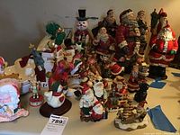 Wide shot showing a collection of various ceramic and resin Santa and snowman figurines with Christmas themes, along with several smaller figurines and decorations on a table.