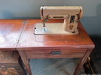 Full view of vintage beige Singer sewing machine mounted into a wooden cabinet with fold-out flaps on either side, closed drawer beneath, wood grain finish and metal hinges.