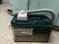 Front side of the green Bissell Little Green Clean Machine showing the clear dirty water tank and attached flexible hose with nozzle.