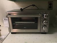 Front view of Oster stainless steel toaster oven showing control dials, glass door, and logo