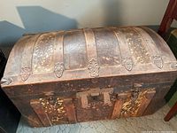 View of side and front of large, antique dome-top trunk with wood, leather and metal construction.