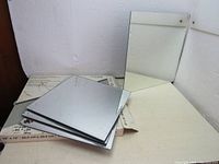 Four 12 inch and three 9.75 inch square mirrors stacked on a surface, original box partially visible, mirrors have polished edges and are in unused condition.
