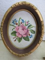 Close-up of one oval cross stitch floral picture in an oval plastic gold frame with glass, showing detailed pink flower pattern and frame condition