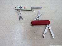 Both pocket tools open showing knife blades and scissors, red Victorinox and white Wenger with Detroit Tigers design