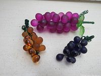 Three vintage glass grape cluster ornaments on a white surface: light purple with green leaf, dark purple without leaf, and amber colored clusters, showing details and sizes.