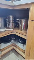 Two open kitchen cabinets showing a stack of shiny stainless steel pots and pans on the top shelf. The lower shelf holds two black cast iron pans and an additional lid.