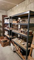 Two metal shelving units with particle board shelves, containing various household items. Located in basement with concrete walls.