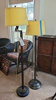 Two bronze tone floor lamps standing near a window with cream lampshades. One lampshade is taped.