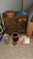 Wooden jewelry chest with six drawers, woven basket on top, two ceramic vases, brass snail figurine and pink soap dish arranged on carpeted floor.