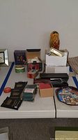 Photo showing multiple items including Russian nesting doll, boxed Kosta Boda bowl, trading card packs, tins, and boxed pocket tool.