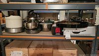 Shelf with stainless steel pots, mixing bowls, plastic salad spinner, 15-inch round metal tray, boxed black non-stick pan with lid.