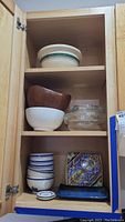 Full cabinet shelf showing various bowls and plates: top shelf white ceramic bowl with green stripe, middle shelf wooden salad bowl and white ceramic bowl with Pyrex glass bowls with lids, bottom shelf stacked Pottery Barn blue striped bowls and a decorative Italian ceramic plate