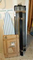 Blue and white striped ironing board, black metal shelving unit parts, and boxed teak bath mat arranged together.