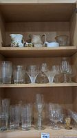 Shelf with assorted glassware including pitchers, vases on top shelf; various drinking glasses, stemware and decorative glass pieces on middle and bottom shelves.