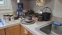 Wide view of kitchen counter showing multiple silverplated and metal items including cups, platters, pots, canisters, French presses, watering can, trivets, and ashtrays