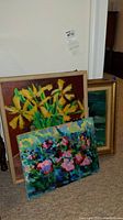 Three original floral paintings, two framed and one unframed, placed on carpeted floor against a white wall. Visible frames on two paintings, one wooden and one gold-colored frame.