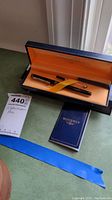 Photo showing Waterman fountain pen in original black and orange lined case with pen secured by orange ribbon; includes a tag labeled 'Waterman Pen' and item number.