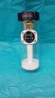 Photo showing white and gold Car Pool Karaoke microphone inside clear plastic cylindrical case with black top labeled 'Car Pool Karaoke The Mic'