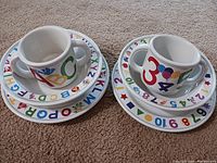 Photo of 2 children's dinner plates, 2 bowls, and 2 cups with colorful alphabet and number designs on the rims and outer sides, arranged on carpet.