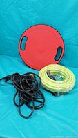 Photo shows a new yellow garden hose coiled and secured, a red circular balance board with black handle cutouts, and a used black extension cord coiled on blue backdrop.