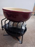 Side view of maroon ceramic fondue pot placed on black metal stand with burner visible underneath and fondue forks inside the pot.